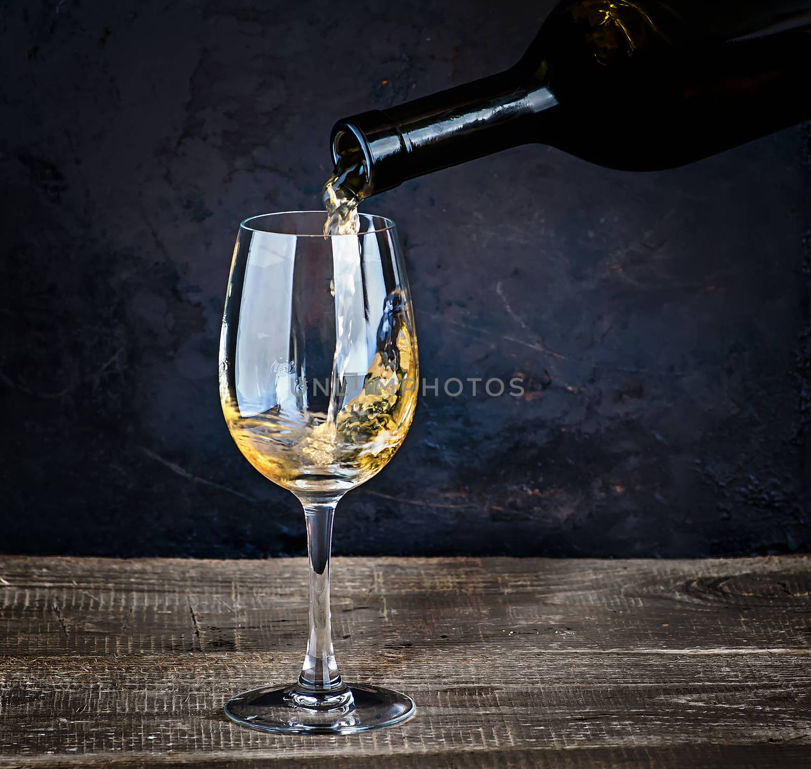 Pouring white wine from bottle into the wineglass on the table on dark blurred background