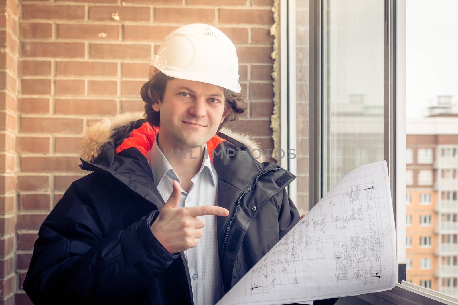 Architect in hardhat, holding blueprints smiling look at the camera and points his finger to the drawing. Soft focus, toned
