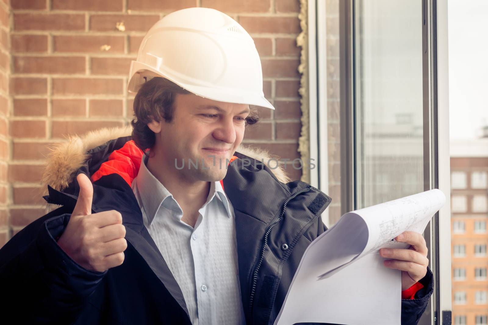 Handsome architect is standing near building outdoors. He is giving thumb up and smiling. The man in helmet is looking at camera happily. Soft focus, toned