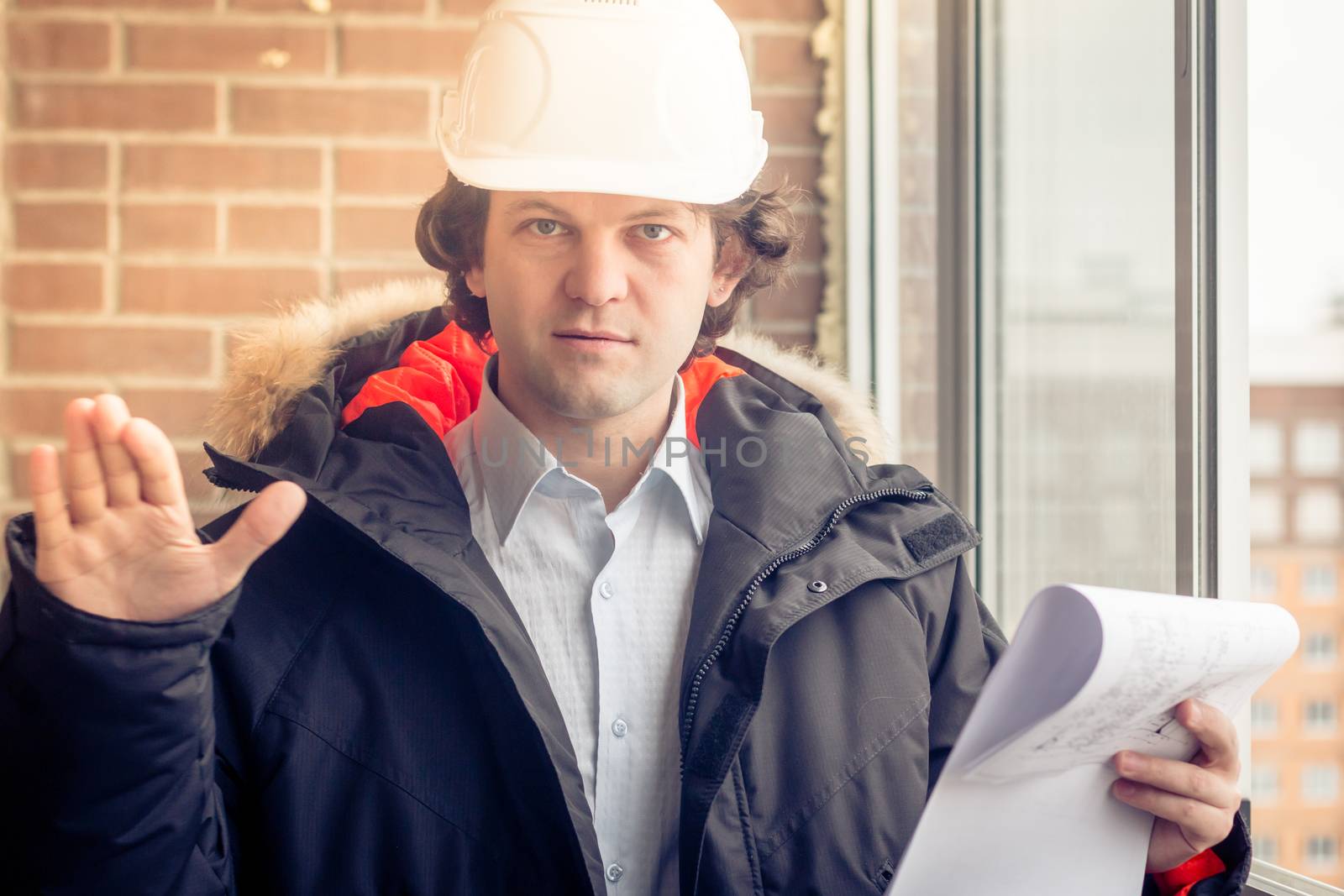 An angry disgruntled builder worker in a helmet with project drawings plans in his one hand and mobile phone in another hand screams on someone. Bad executed work. Aggressive man. Soft focus, toned