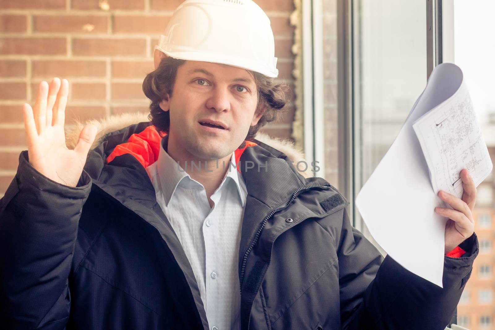 An angry disgruntled builder worker in a helmet with project drawings plans in his one hand and mobile phone in another hand screams on someone. Bad executed work. Man in panic. Soft focus, toned