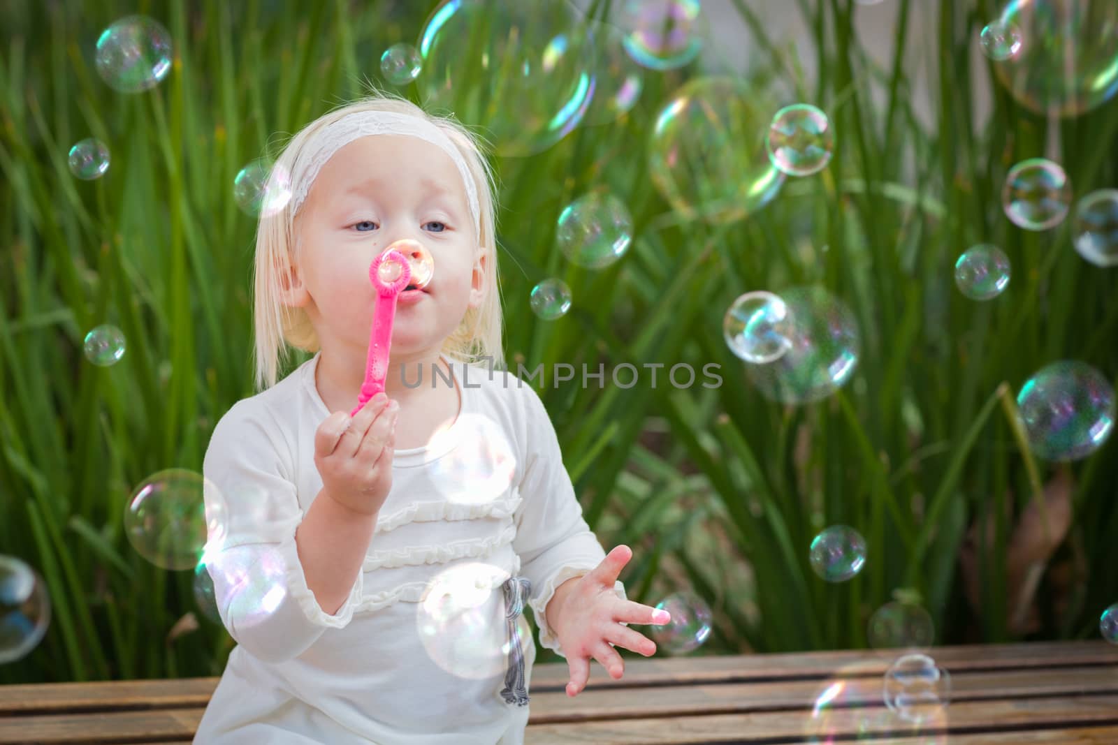 Adorable Little Girl Sitting On Bench Having Fun With Blowing Bubbles Outside. by Feverpitched