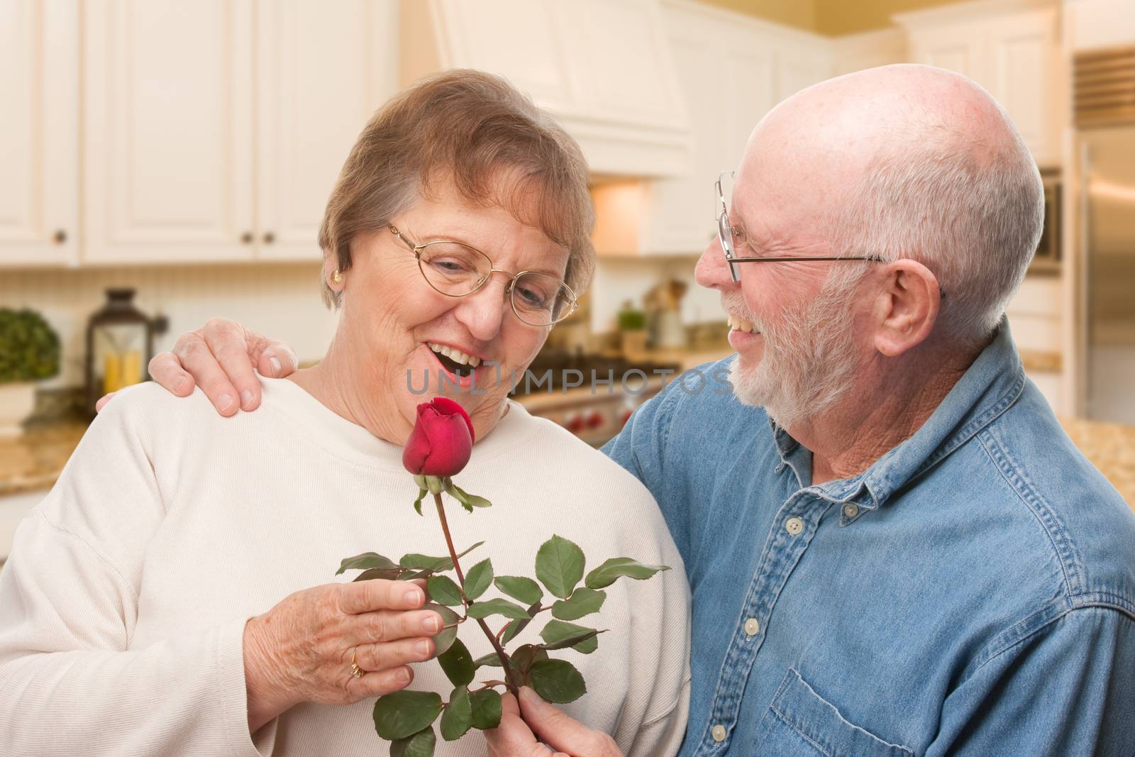Happy Senior Adult Man Giving Red Rose to His Wife Inside Kitchen. by Feverpitched