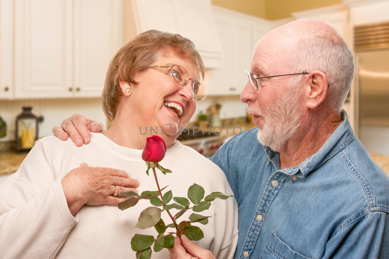 Happy Senior Adult Man Giving Red Rose to His Wife Inside Kitchen. by Feverpitched