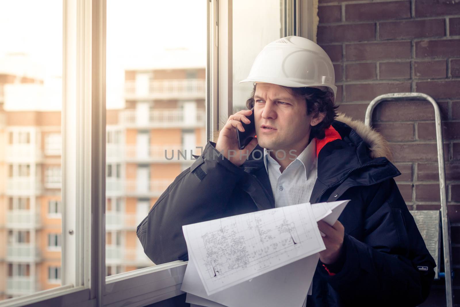 Portrait of concerned unshaven builder telling by mobile while keeping different projects in hand. Communication and work concept. Soft focus, toned. by MSharova