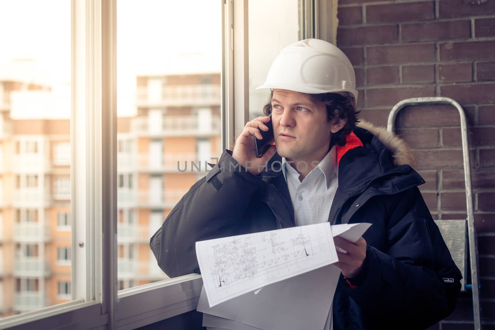 Portrait of concerned unshaven builder telling by mobile while keeping different projects in hand. Communication and work concept. Soft focus, toned. by MSharova