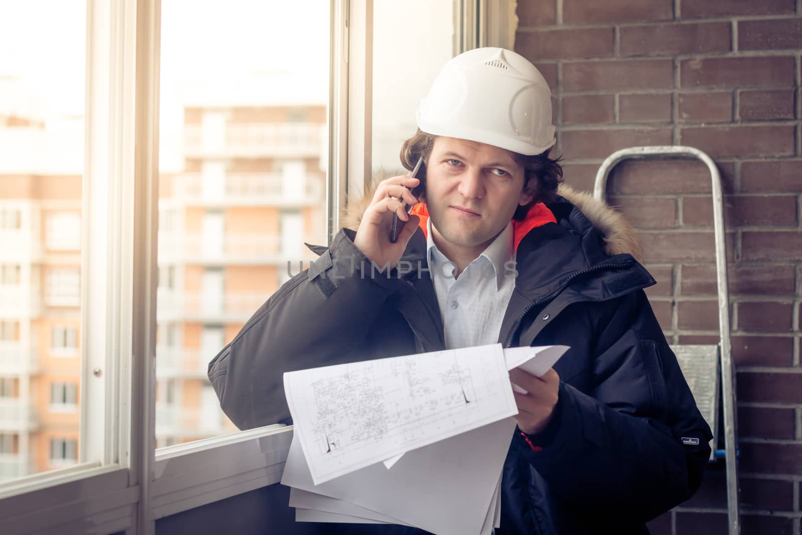 Portrait of concerned unshaven builder telling by mobile while keeping different projects in hand. Communication and work concept. Soft focus, toned