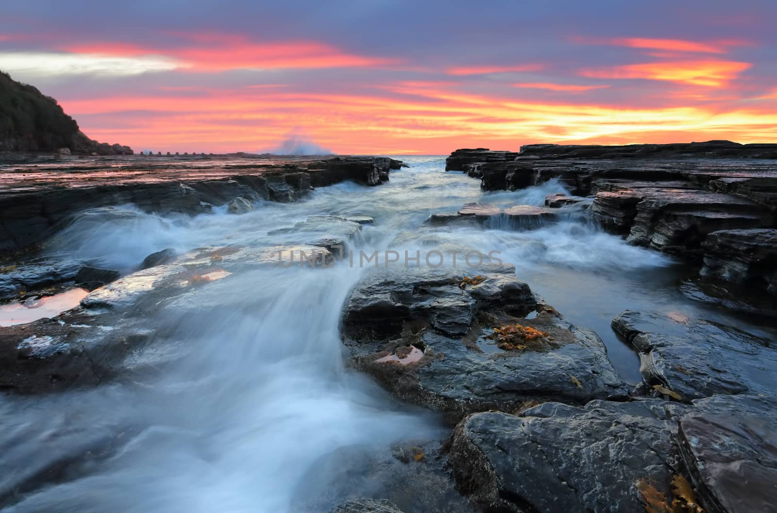  Beautiful sunrise lights up the clouds as waves flow into eroded rock chasm flowing over and around loose rocks