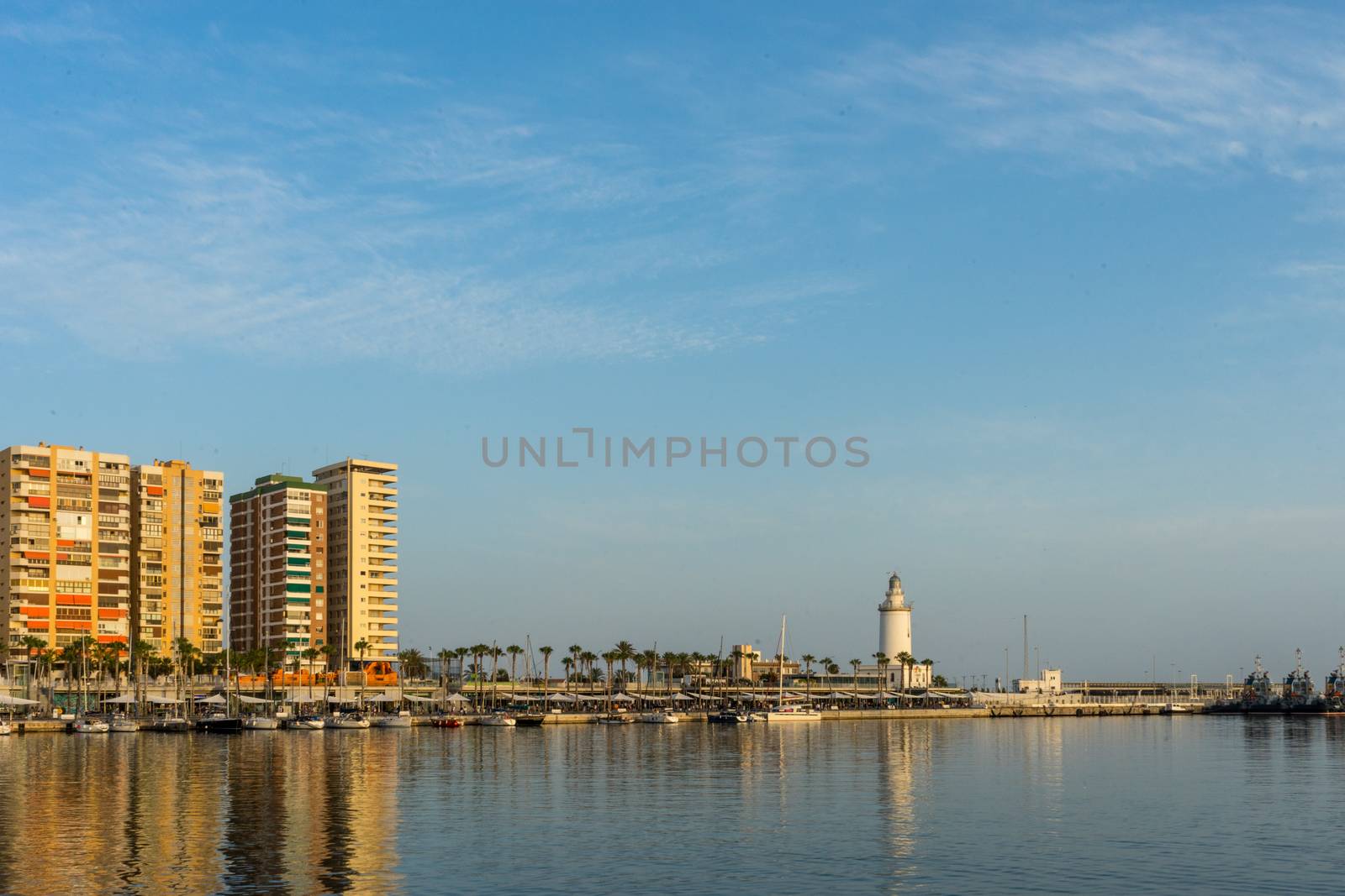 White lighthouse and the tall buildings of Malaga with their reflections in Malaga, Spain, Europe at golden sunset hour