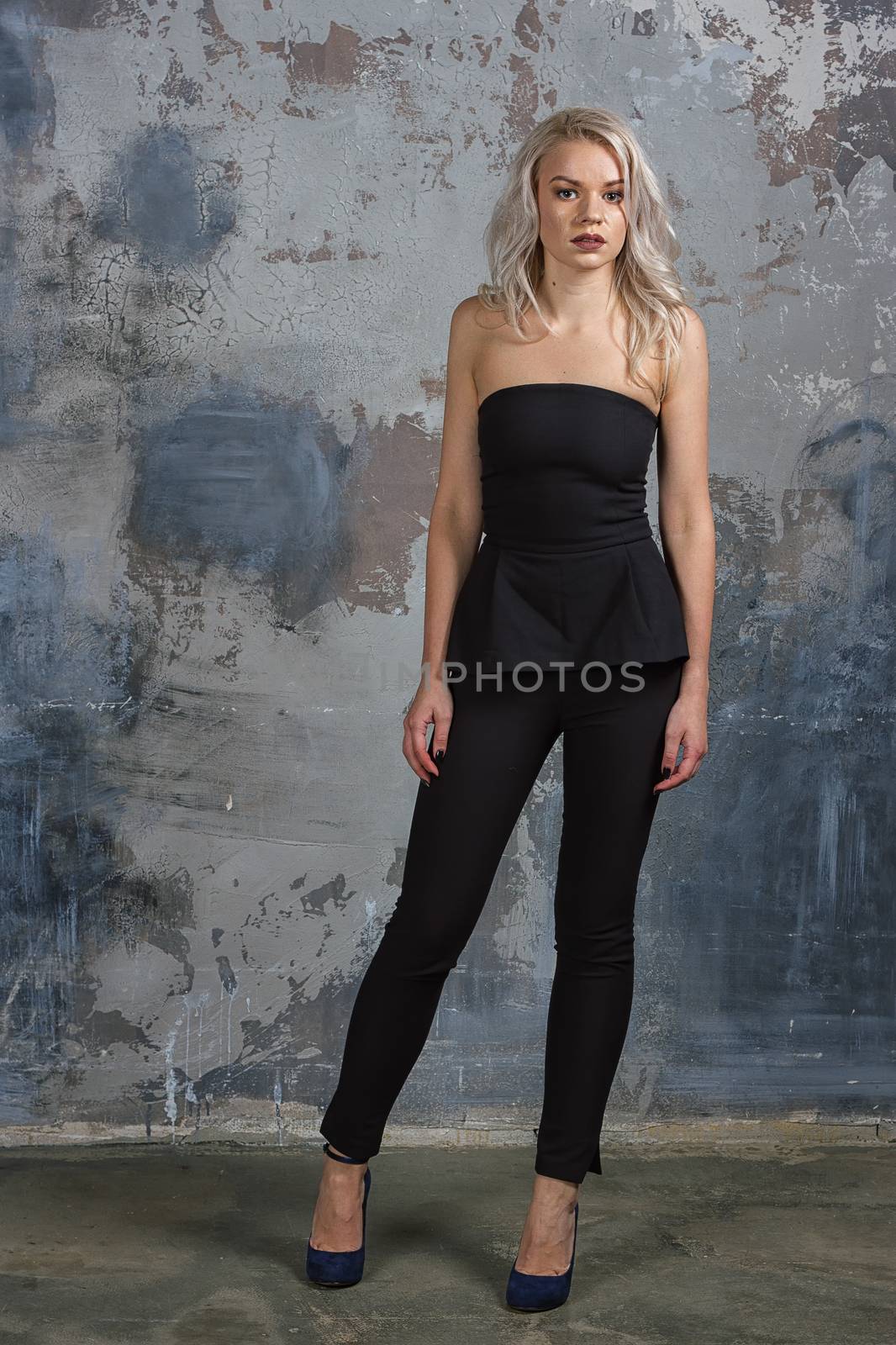 Young beautiful pretty woman with long curly hair and red lips in black overall standing against wall and looking at you. Fashionable caucasian girl posing in simple interior
