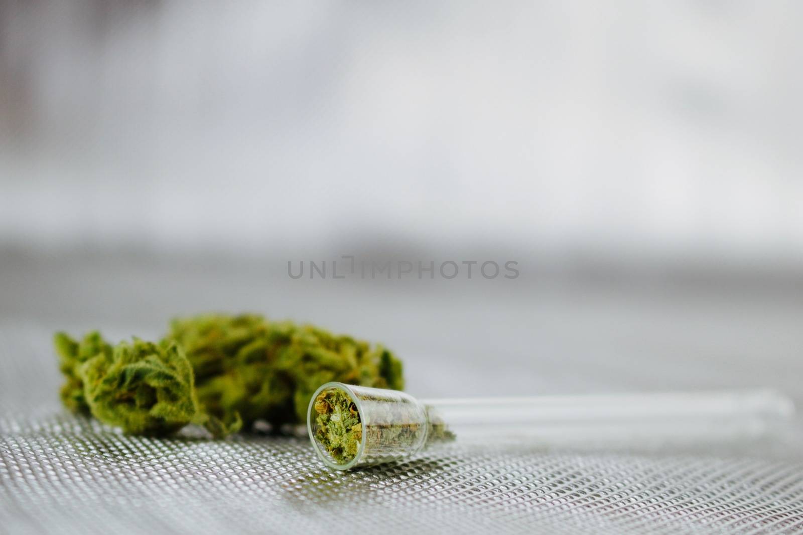 Transparent small glass pipe and dry green marijuana leafs on silver background.