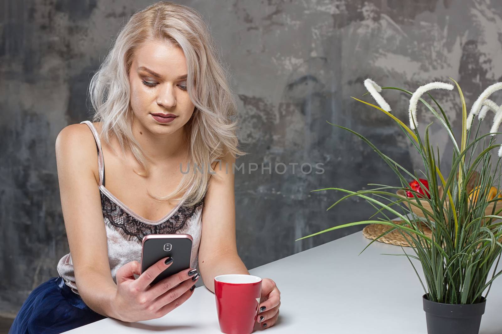 Beautiful blonde girl in home clothes is using a smartphone and smiling while sitting in kitchen