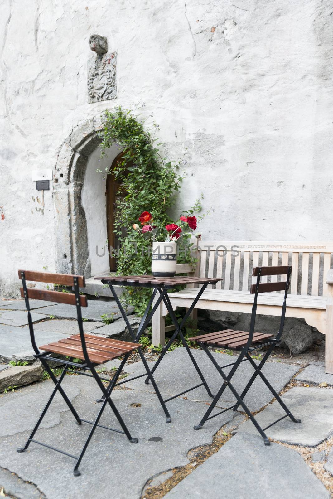 old terrace in bergen norway with red flowers and table and chairs in front of old wall with bow entrance