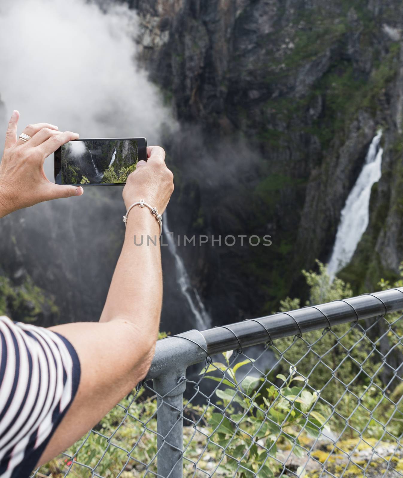 woman making photo with mobile phone of the Voringfossen waterfall in norway seen from street level on sunny day 