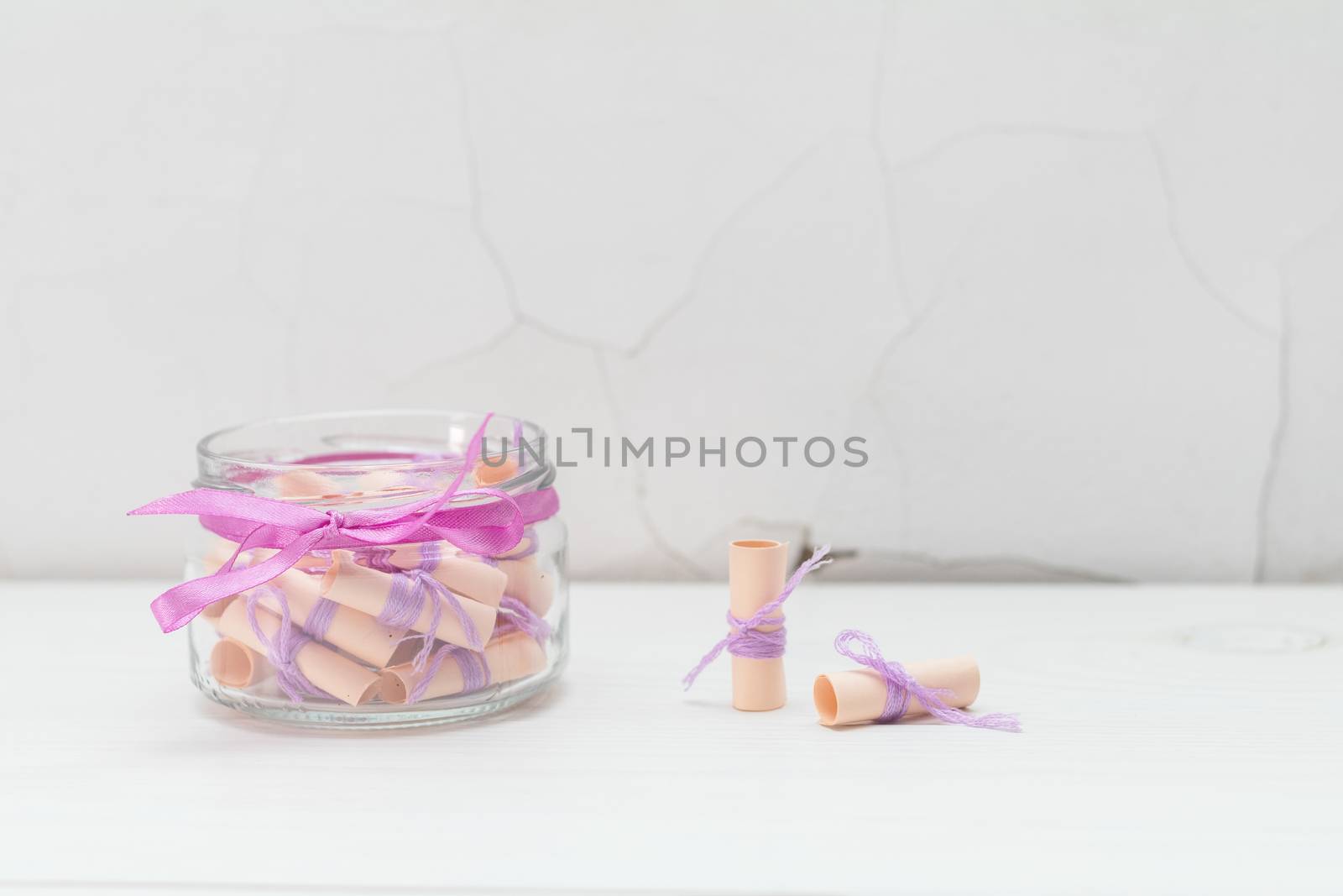 Paper wish scrolls witj violet threads in a glass jar with violet ribbon. White wooden surface close to the white cracked wall. Light tones, copy space.