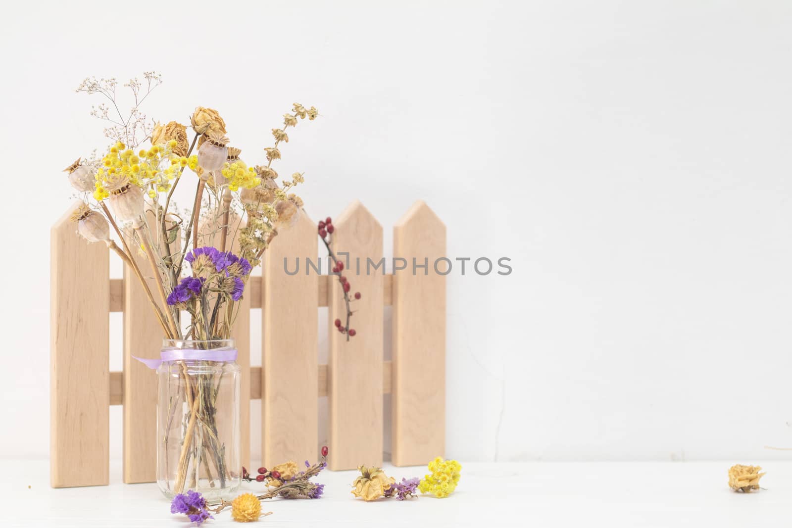 Dried flowers and poppy heads on a stem in a glass jar. Wooden decoration. White wooden table, white cracked wall. Light tones, copy space.