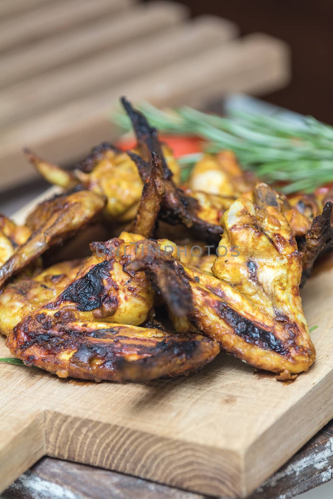 Pile of roasted grilled chicken wings on oak wooden board. Fresh vegetables and greens on the background. Fresh grilled chicken meat. Shallow depth of field.
