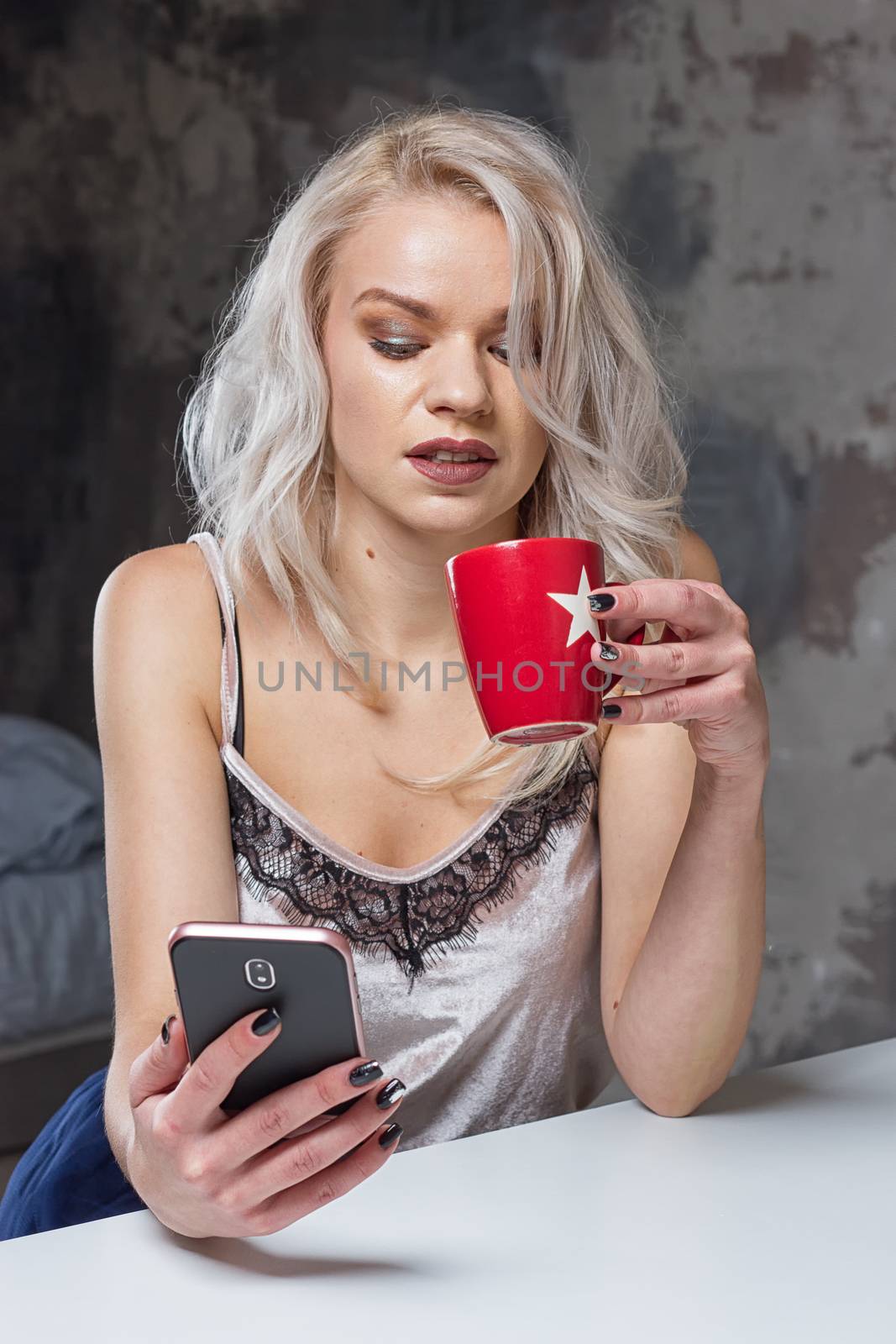 Beautiful blonde girl in home clothes is using a smartphone and smiling while sitting in kitchen