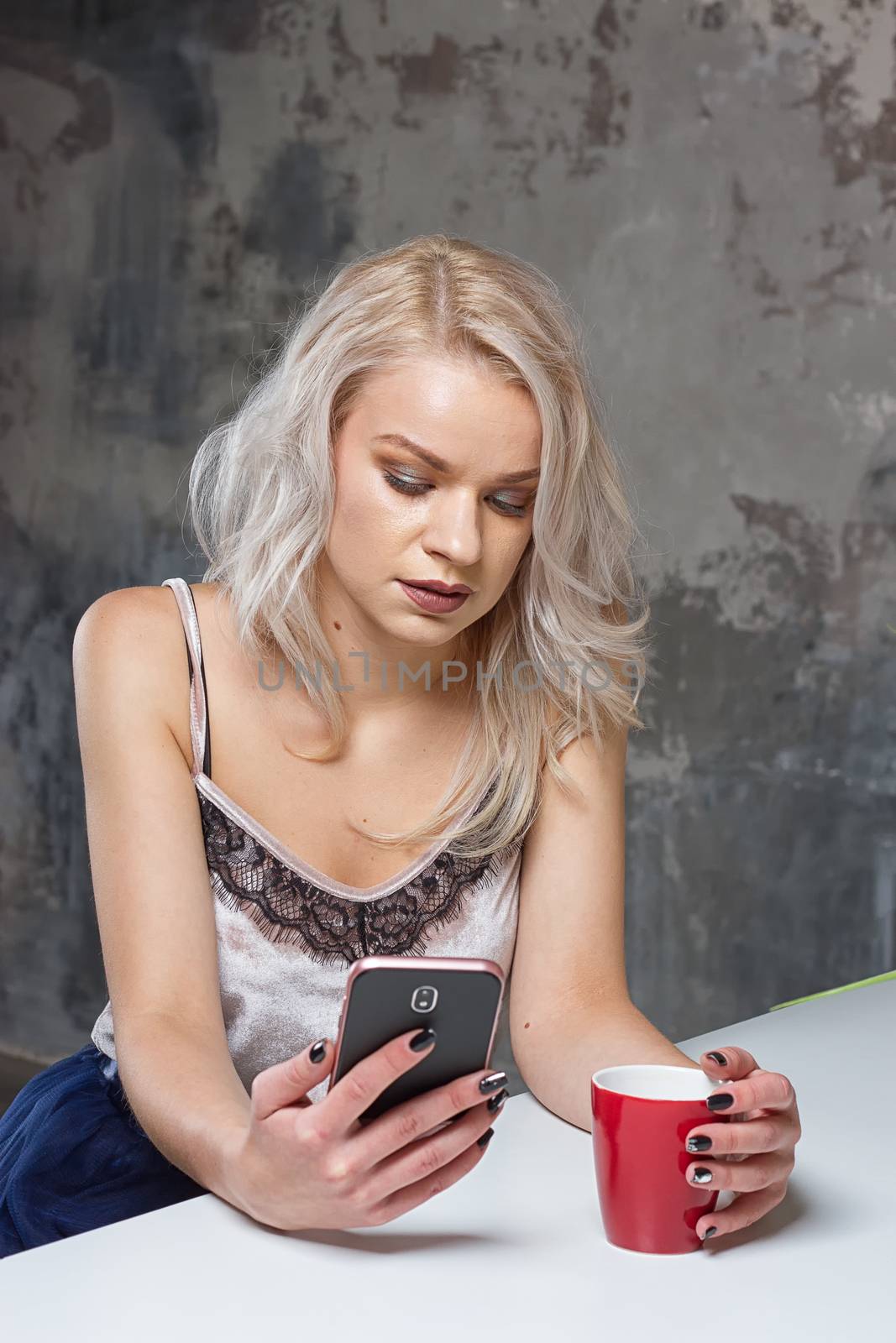 Beautiful blonde girl in home clothes is using a smartphone and smiling while sitting in kitchen