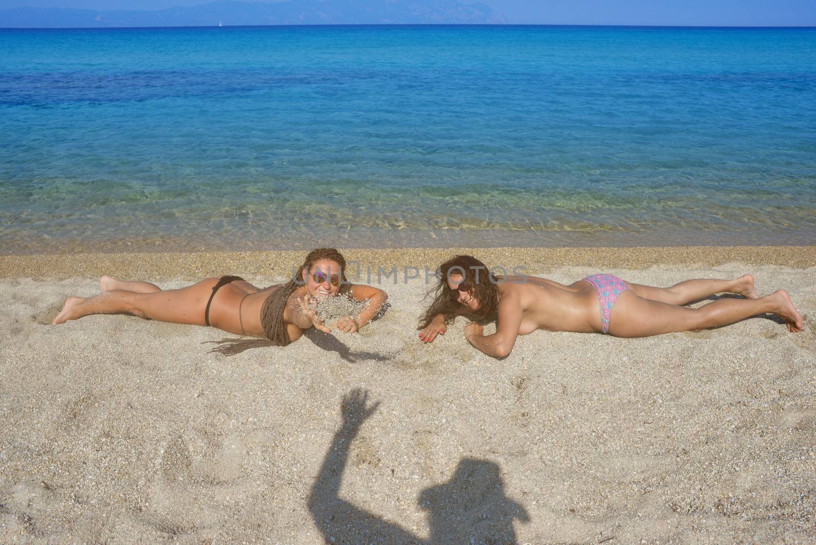 Two beautiful women on the beach are smiling to a person, shown with his waving shadow on the sand.