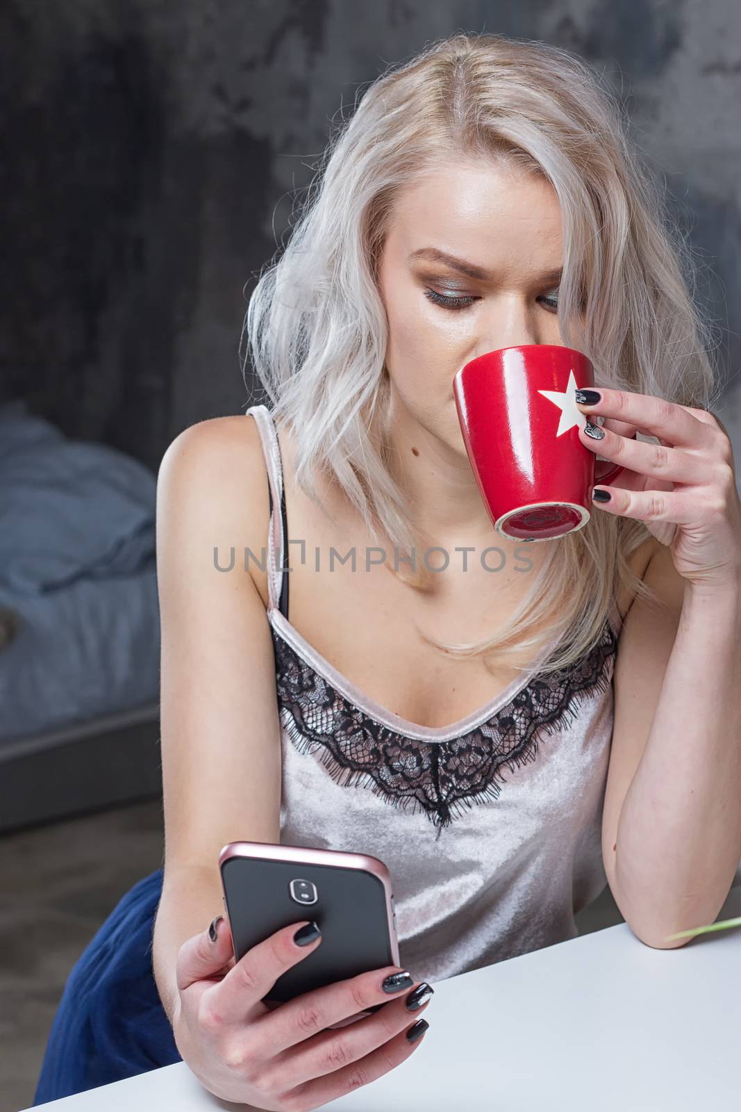 Beautiful blonde girl in home clothes is using a smartphone and smiling while sitting in kitchen