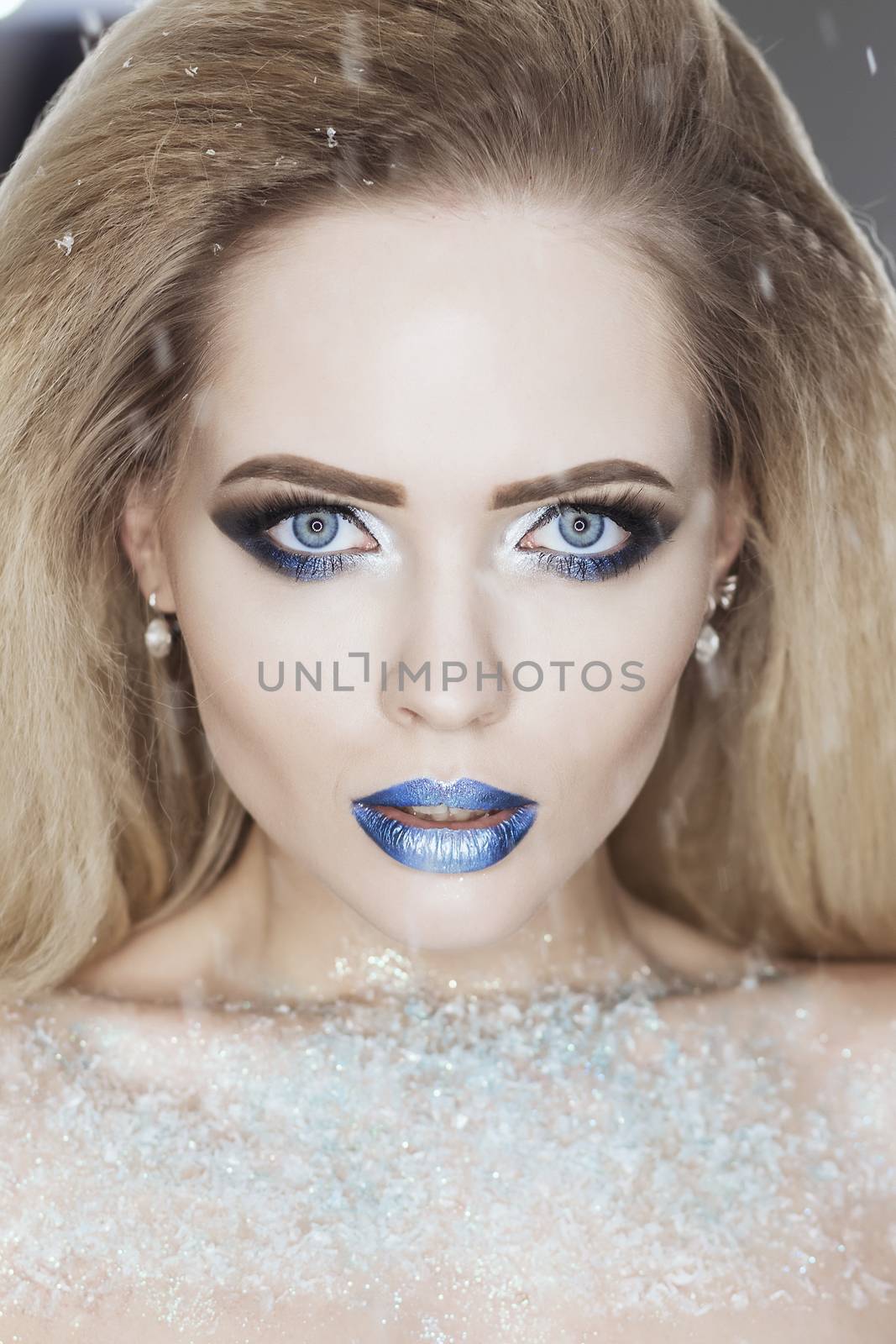 Winter Beauty Woman. Christmas Girl Makeup. Holiday Make-up. Snow Queen High Fashion Portrait over Blue Snow Background. Eyeshadows, False Eyelashes and Crystals on the Lips. Copy Space for Your Text.