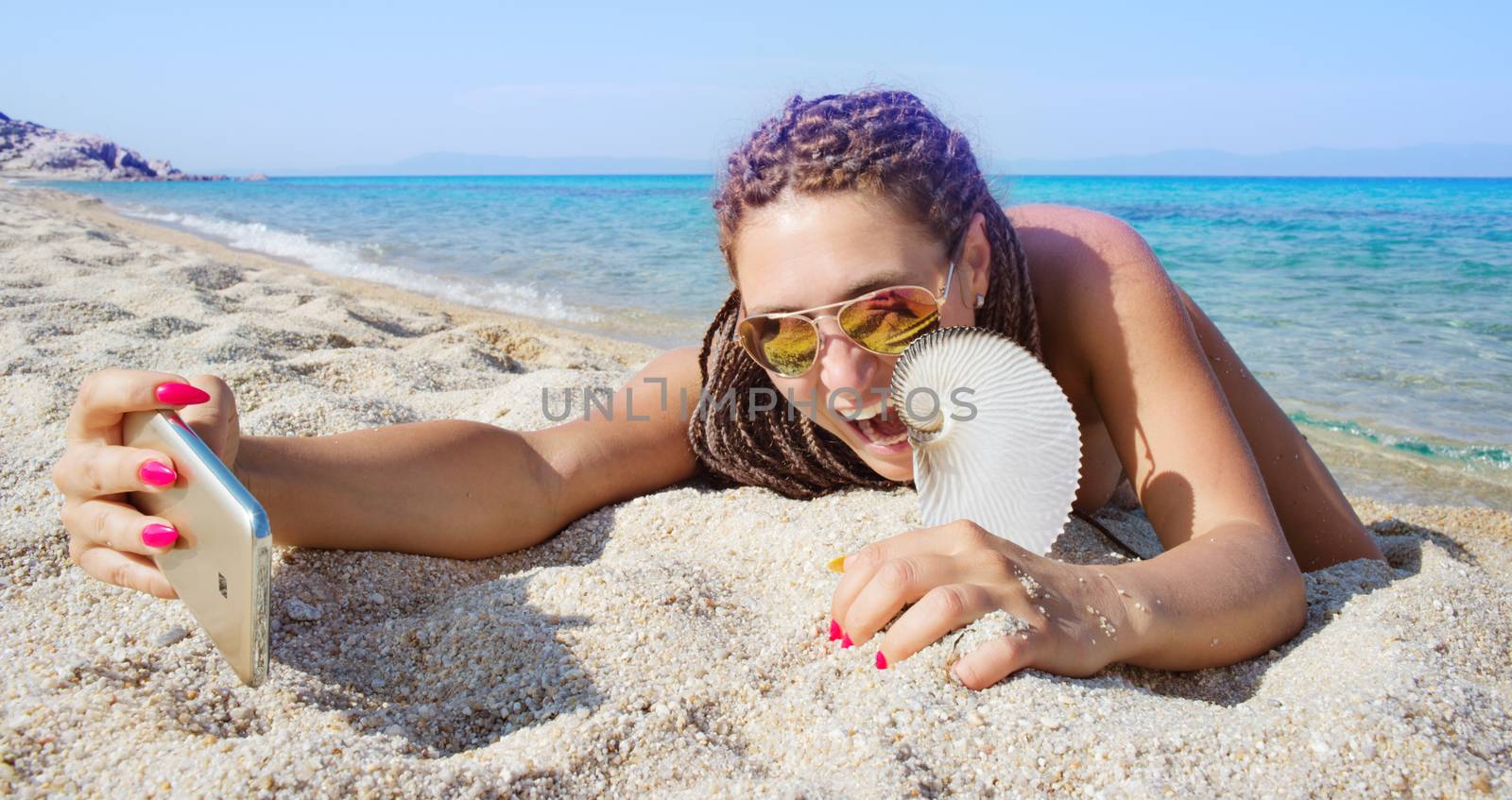 Gorgeous female is making selfie with smartphone lying on a sandy beach of a beautiful distant sea shore, holding nautilus shell.