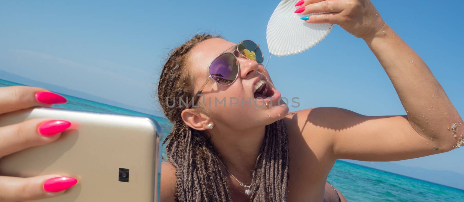 Beautiful female is making selfie on a remote beach while drinking water out of a nautilus shell.