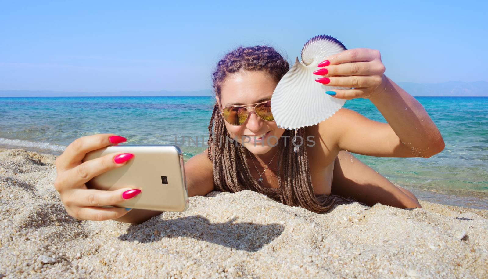 Gorgeous female is making selfie with smartphone lying on a sandy beach of a beautiful distant sea shore, holding nautilus shell.