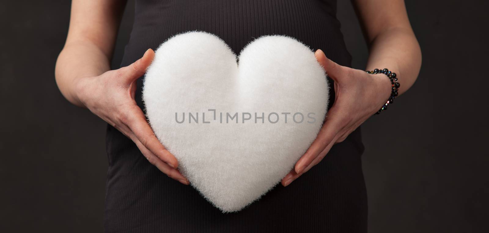 Hands of pregnant woman hold a white heart in front of her abdomen.