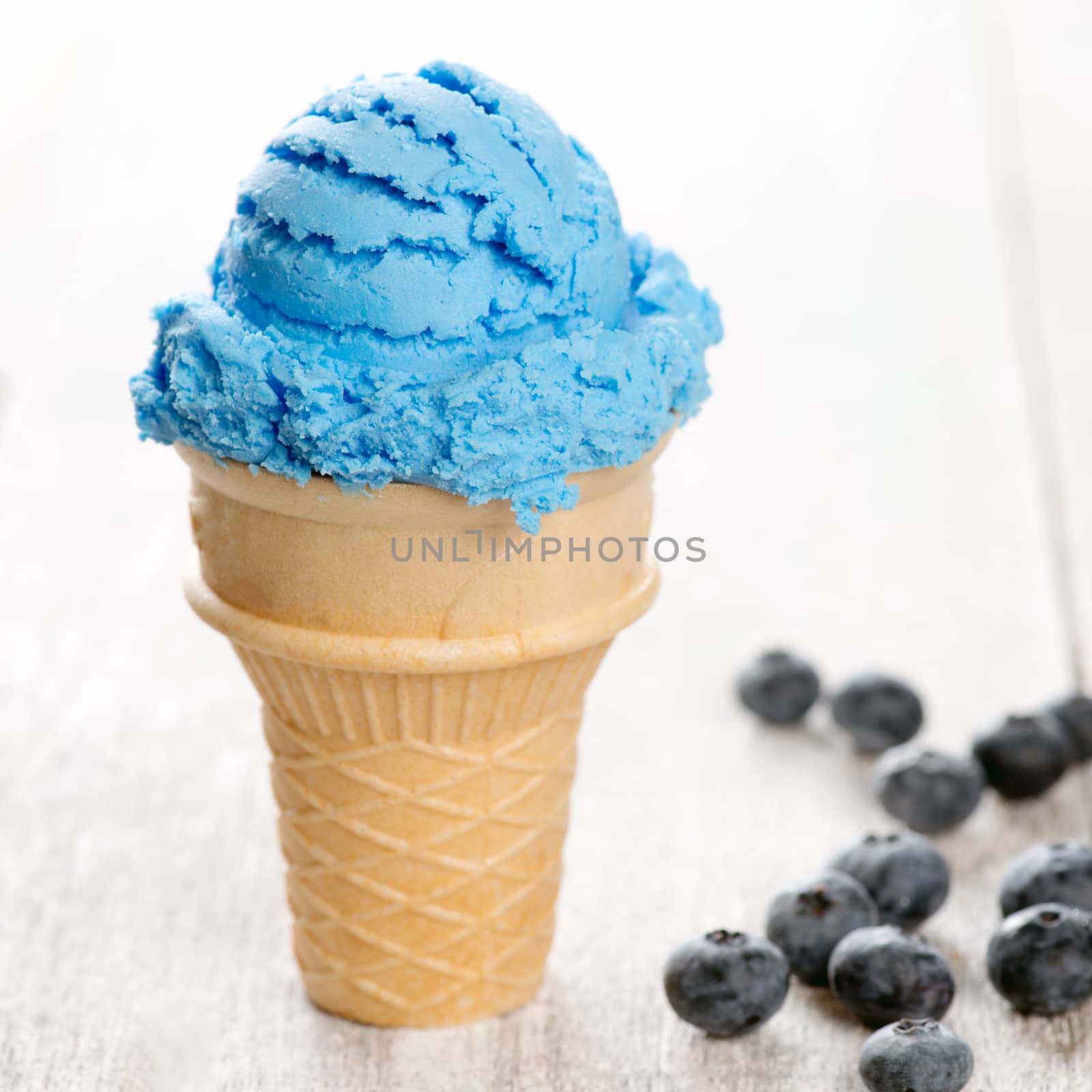 Blue ice cream in waffle cone with blueberry fruit on white rustic wooden background.