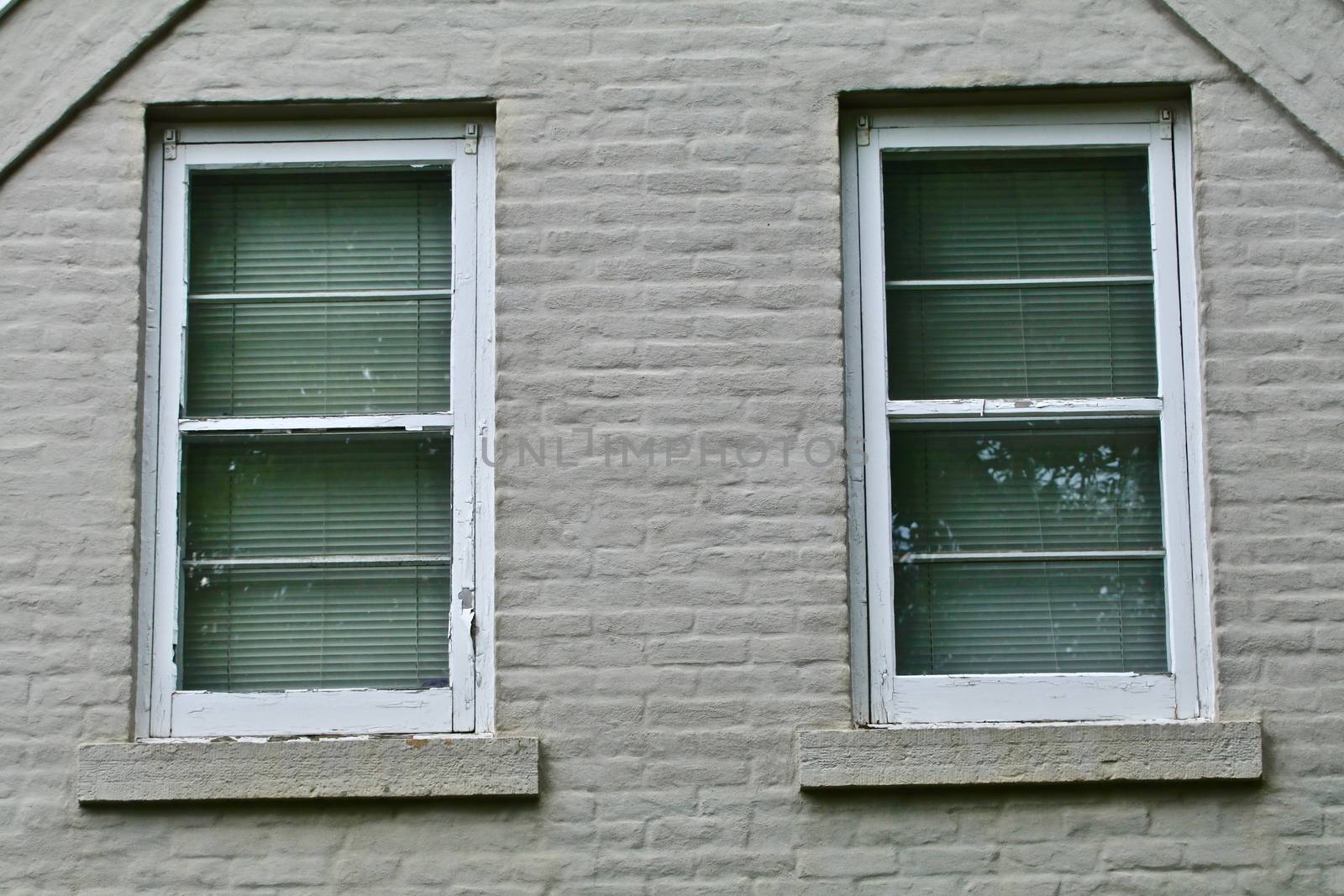 A pair of white windows appear side by side in an old gray painted brick house.
