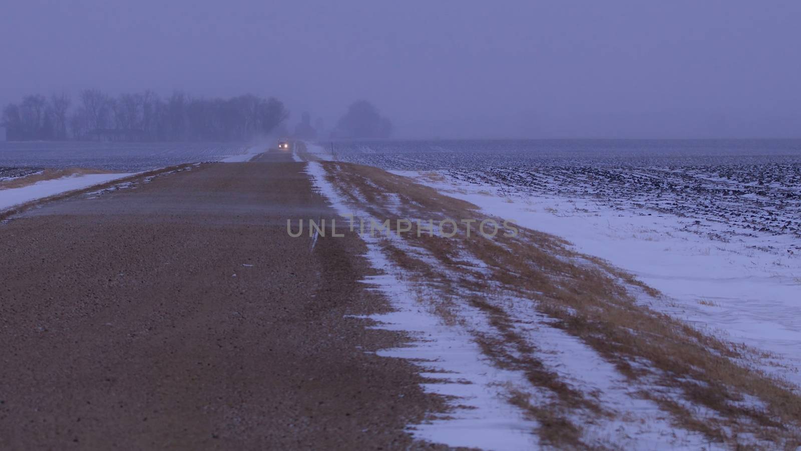 A car comes from the distance down a rural road in a blizzard.