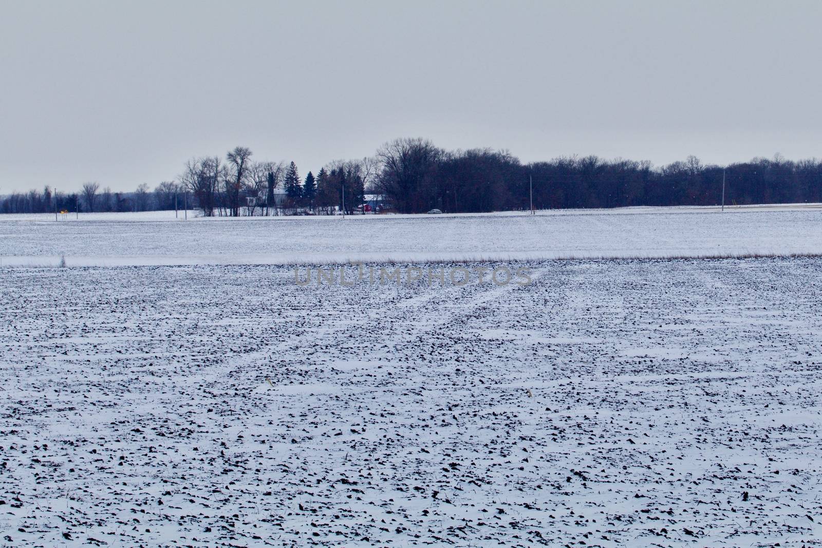 A farmsted in the background with plowed fields in the foreground covered with white snow shows.