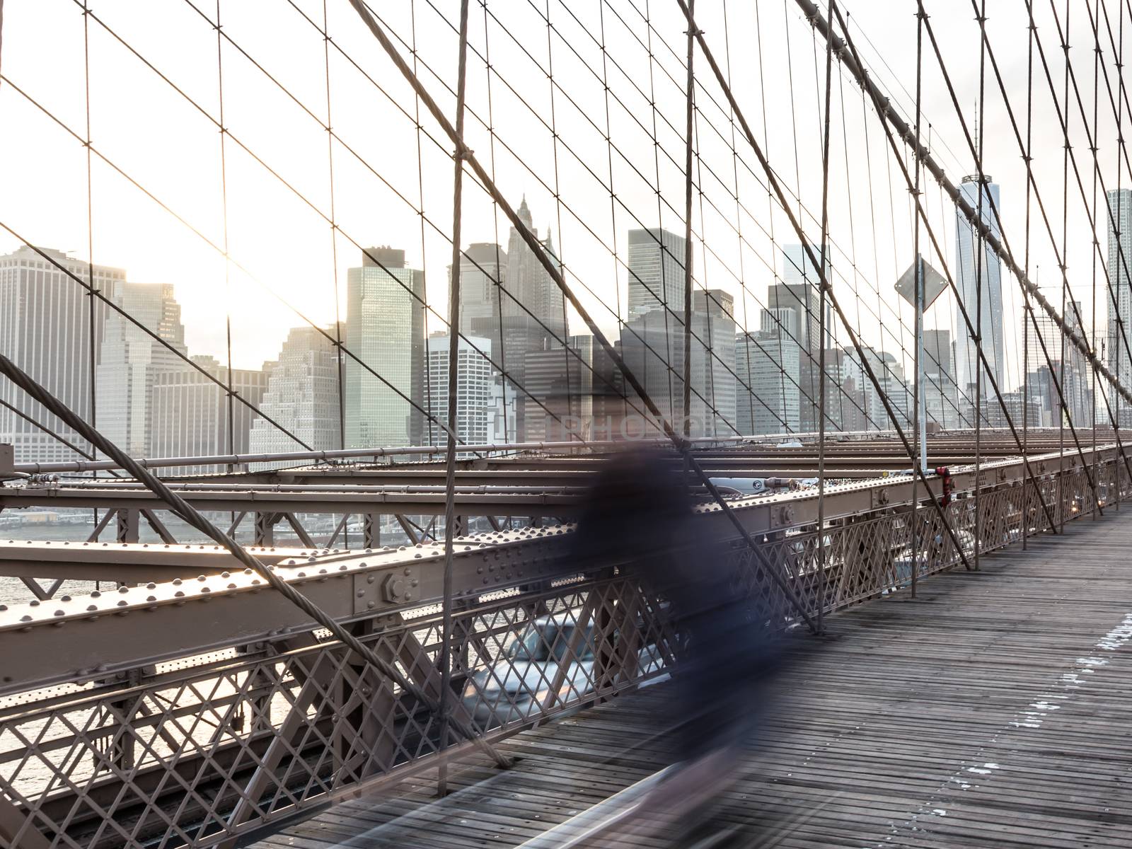 Blured cyclist cycling across Brooklyn bridge. New York City Manhattan downtown skyline in sunset with skyscrapers illuminated over East River panorama as seen from Brooklyn bridge.