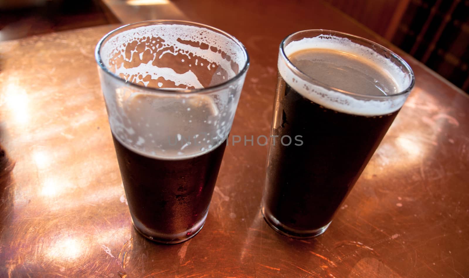 2 tall glasses of dark beer on a copper top table by experiencesnw