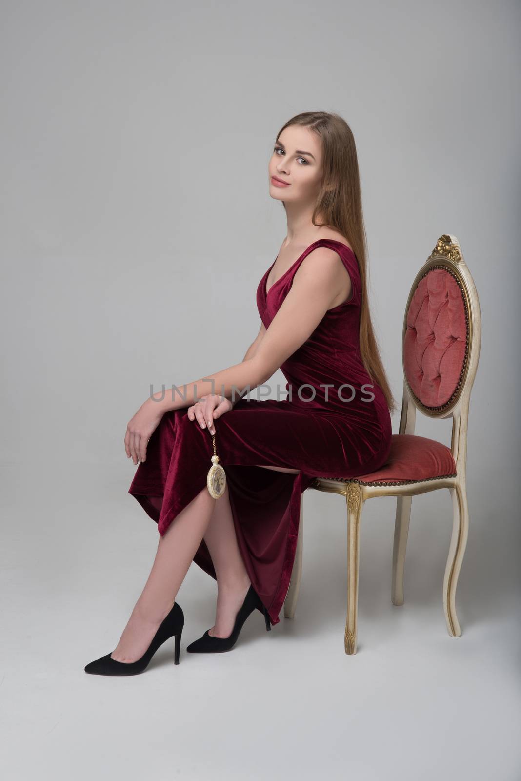 Young beautiful long-haired female model poses in long red dress near red chair on grey background