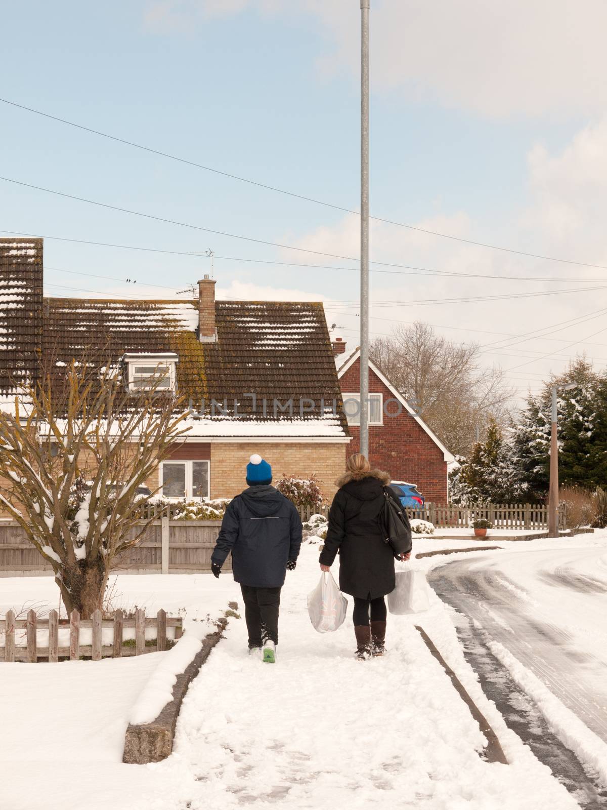 mother and son walking down street with snow in winter uk estate from behind; essex; england; uk