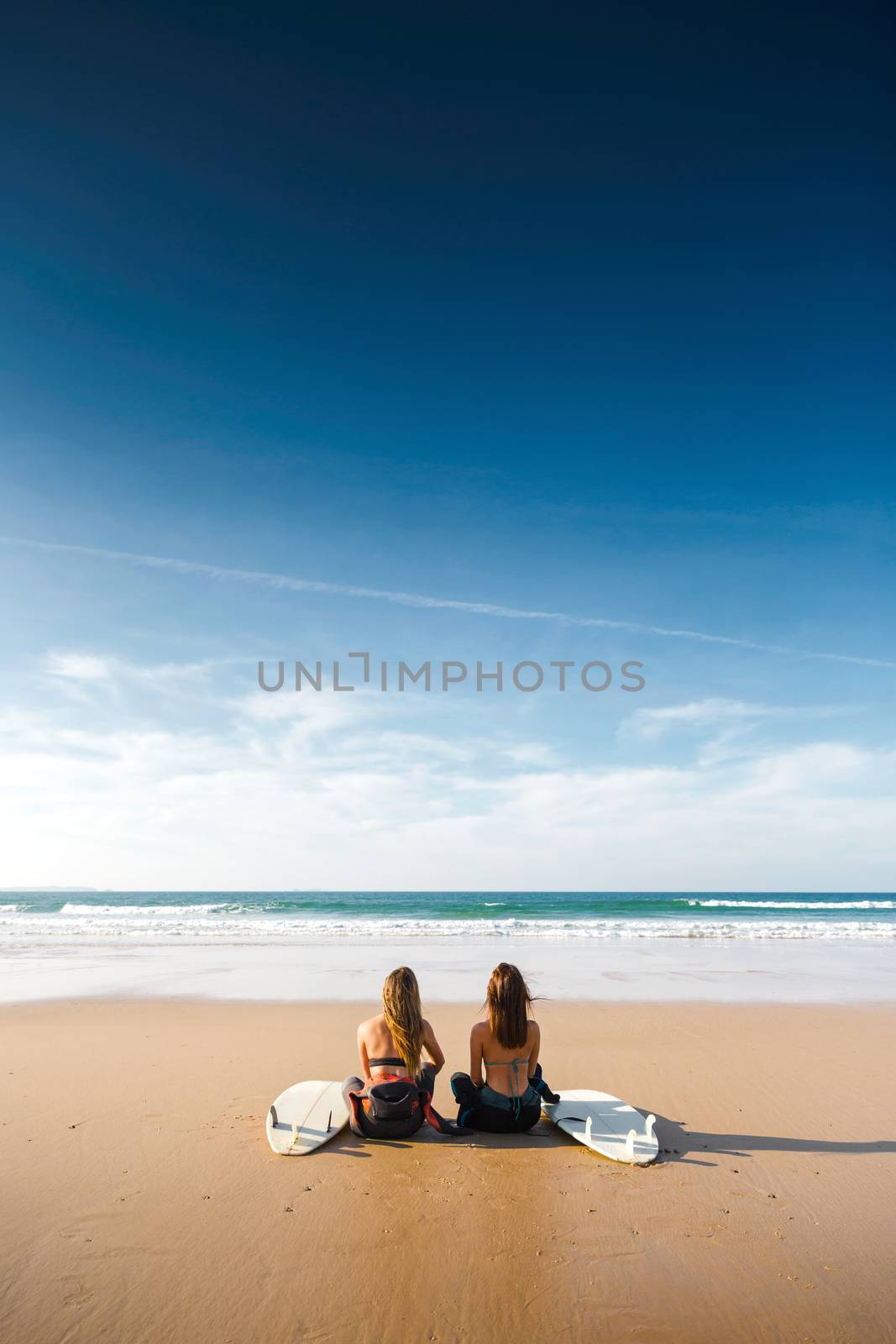 Wide view of two surfer girls on the beach with her surfboards 
