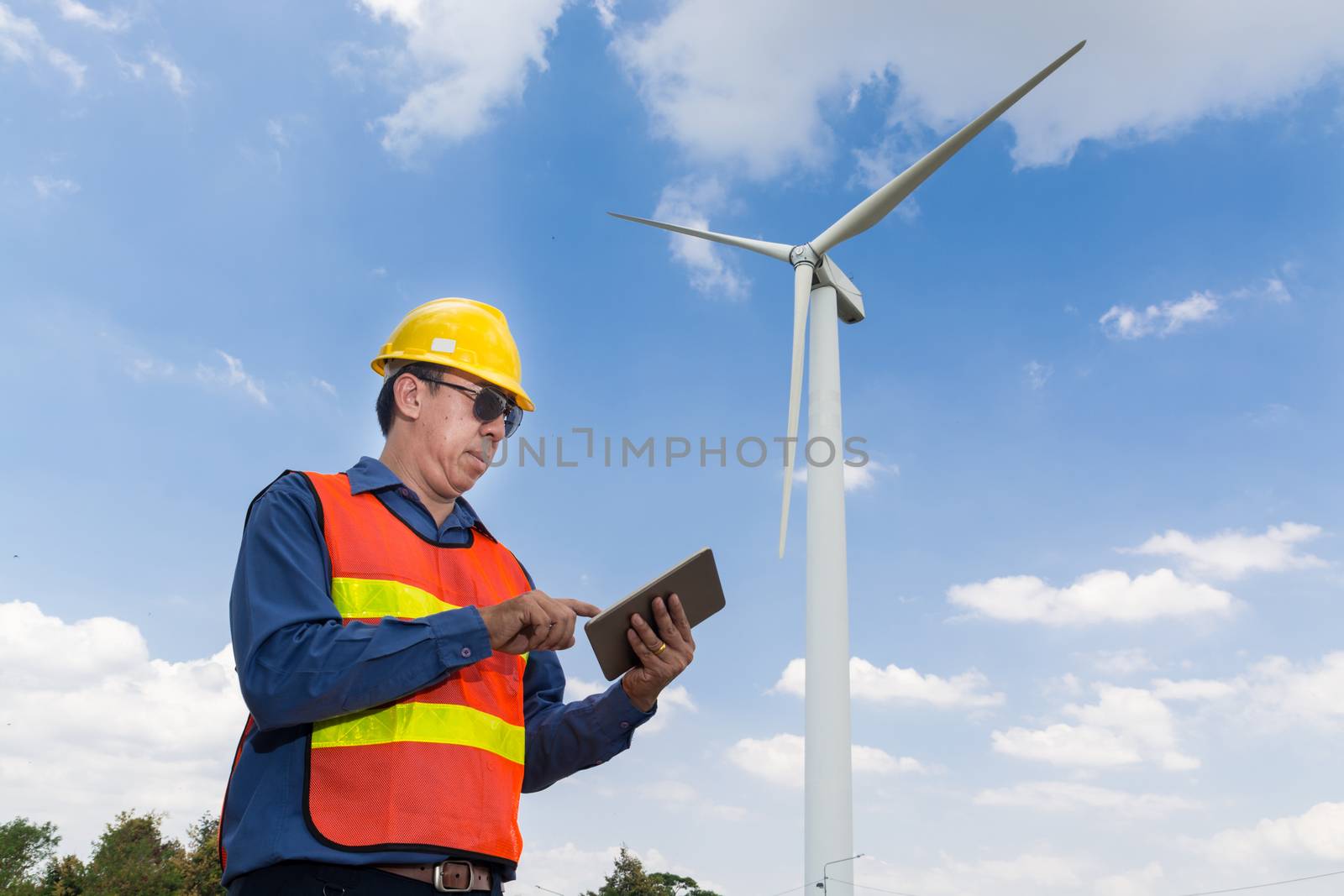 Electric Engineer use Digital Wireless Tablet Device with Wind turbine power Generator Tower Background as Green energy or Renewable Energy Technology Project Development Concept.