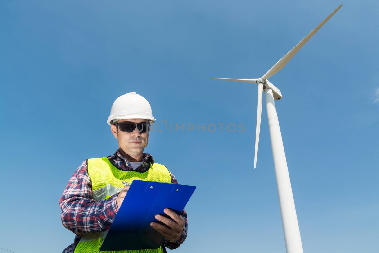 Electric Engineer writing Maintenance report on Clipboard with Wind turbine power Generator Tower Background as Green energy or Renewable Energy Technology Project Development Concept.