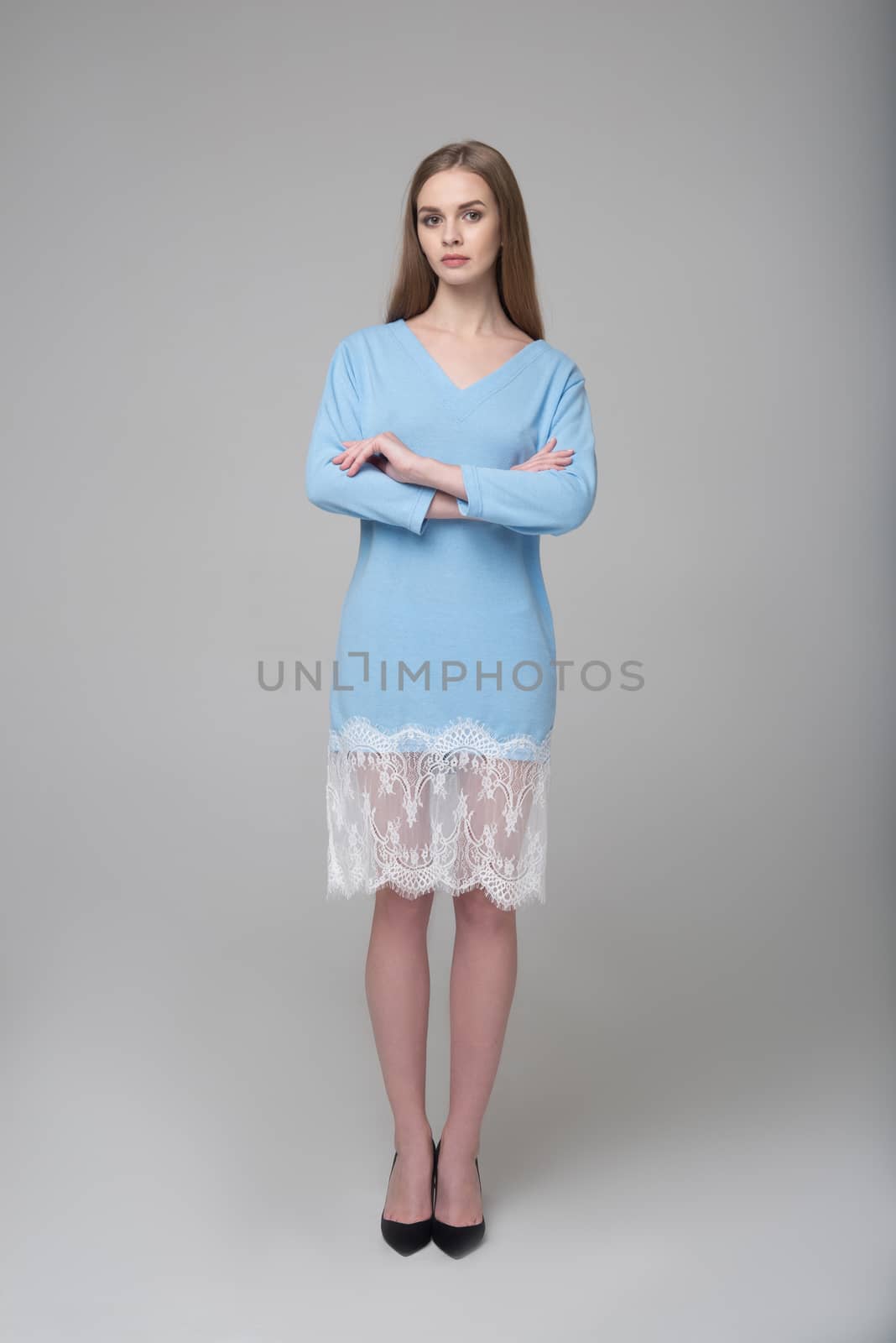 Young beautiful long-haired female model poses in blue dress with white lace on grey background