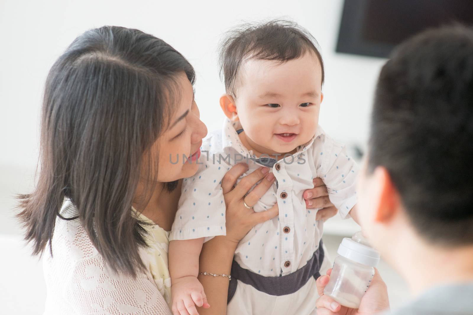 Daddy showing milk bottle to baby. Happy Asian family at home, candid living lifestyle indoors.