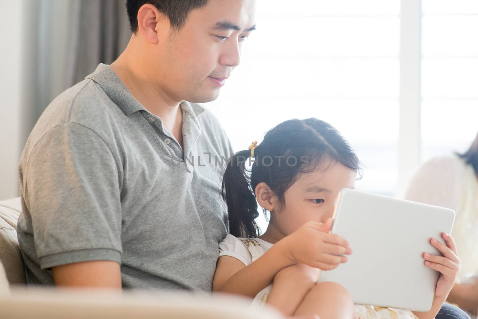 Father using touch screen tablet PC with daughter on sofa. Asian family at home, living lifestyle indoors.