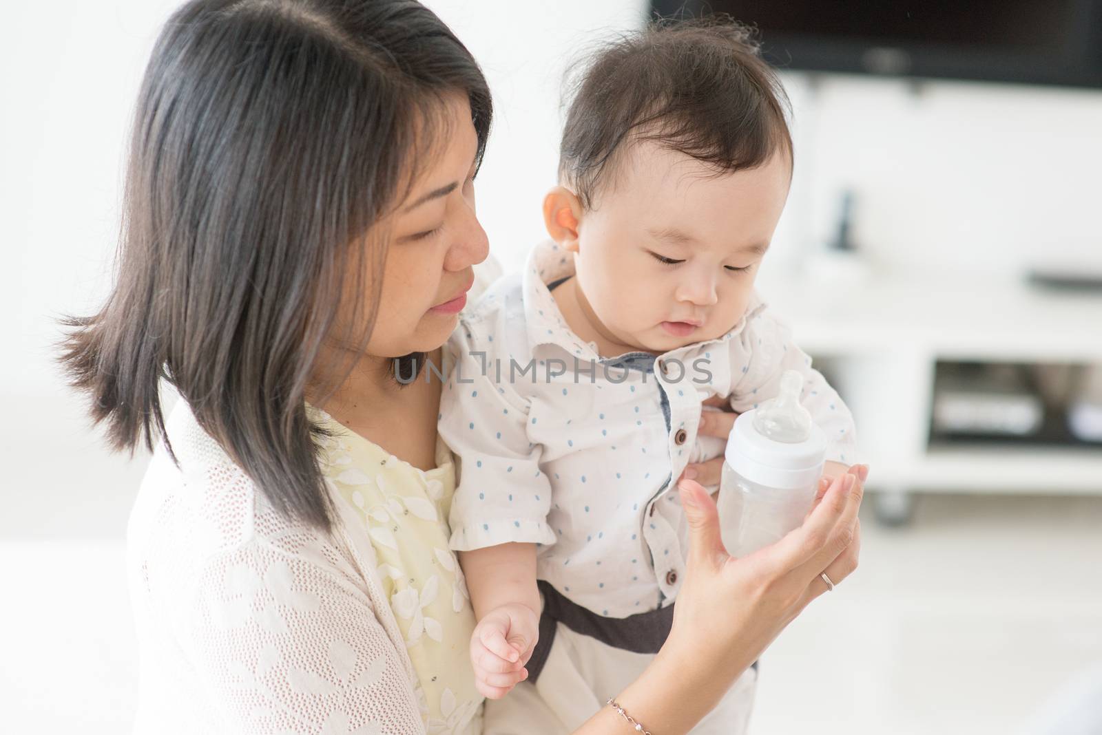 Mother holding milk bottle with 9 months old baby. Asian family at home, living lifestyle indoors.