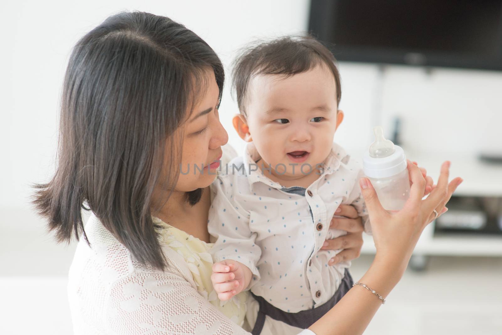 Mother holding milk bottle with 9 months old child. Asian family at home, living lifestyle indoors.