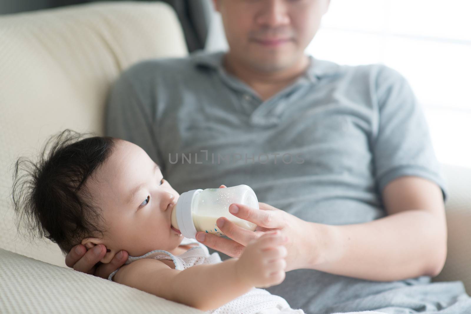 Happy Asian family at home. Father bottle feed formula milk to 9 months old child, living lifestyle indoors. 