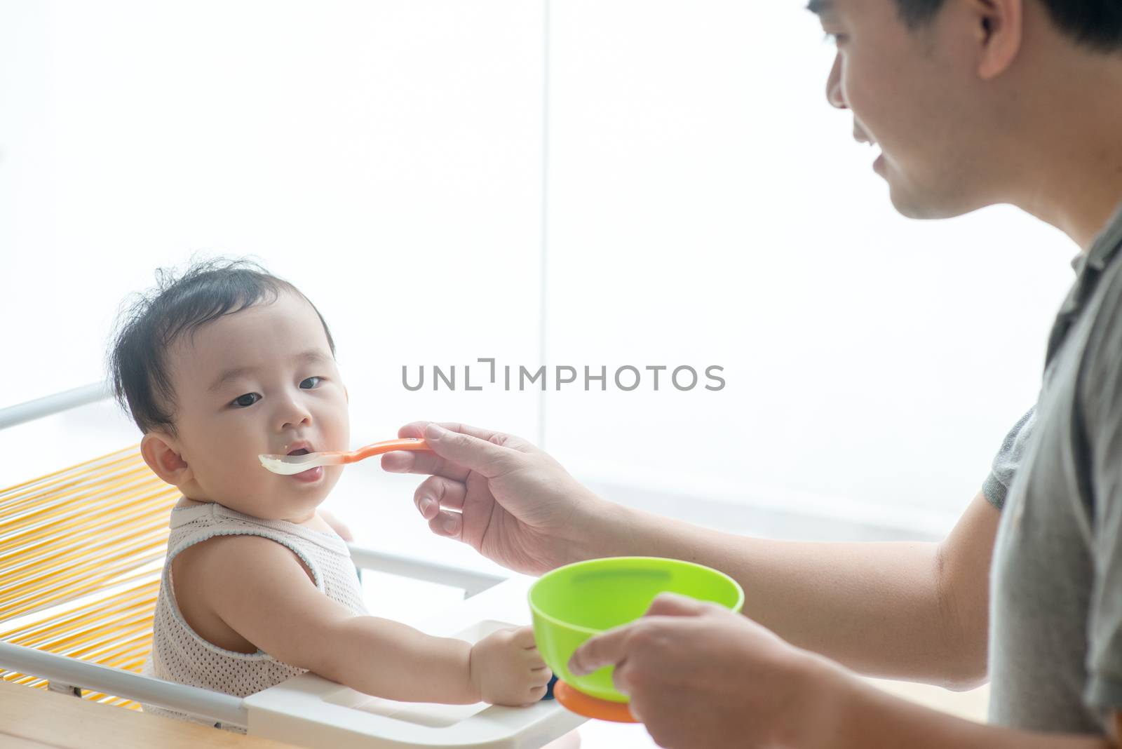 Happy Asian family at home. Father feeding solid food to his 9 months old baby boy in the kitchen, living lifestyle indoors. 