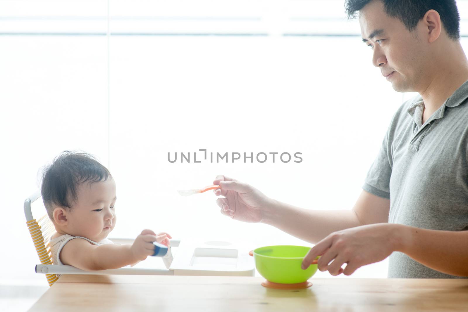Happy Asian family at home. Father feeding solid food to his 9 months old toddler in the kitchen, living lifestyle indoors. 