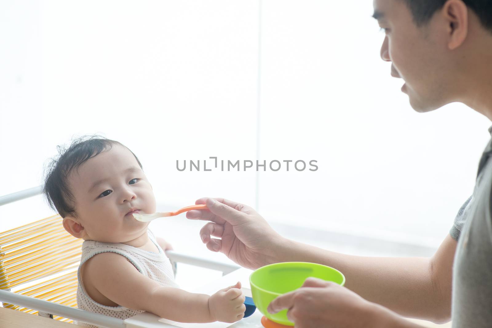 Happy Asian family at home. Father feeding solid food to his 9 months old baby in the kitchen, living lifestyle indoors. 